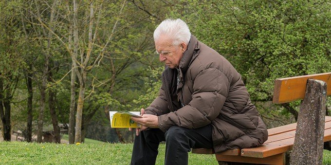 Äldre man sitter på en parkbänk och läser en bok.